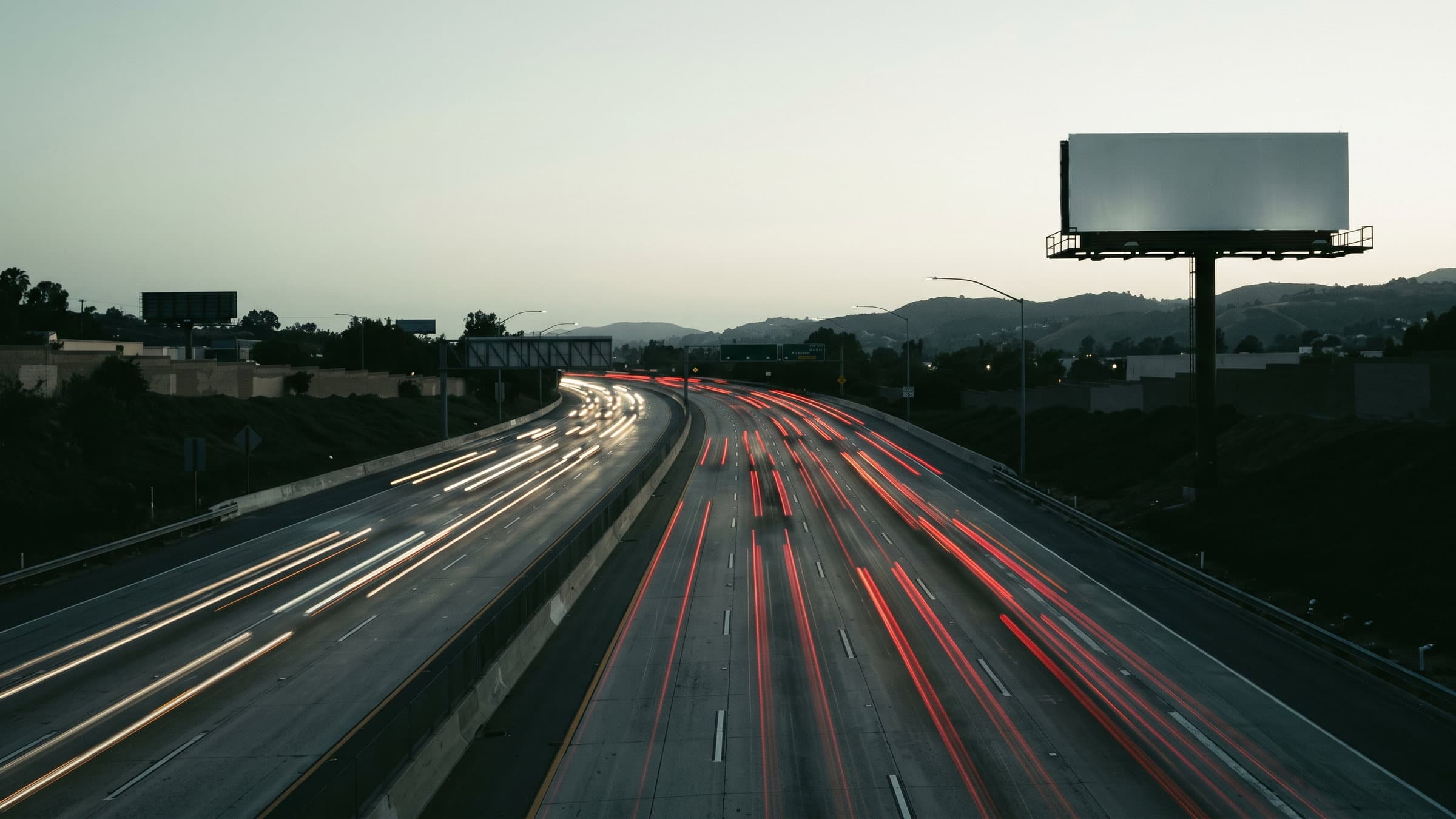 A California freeway at dusk seen from an overpass, with an empty, unlit billboard frame on the shoulder.
